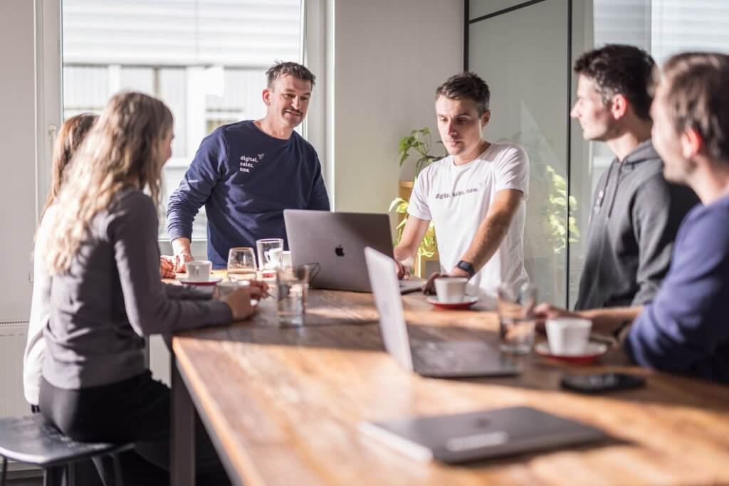 Teammeeting in modernem Büro, Personen diskutieren am Tisch mit Laptops und Kaffeetassen.